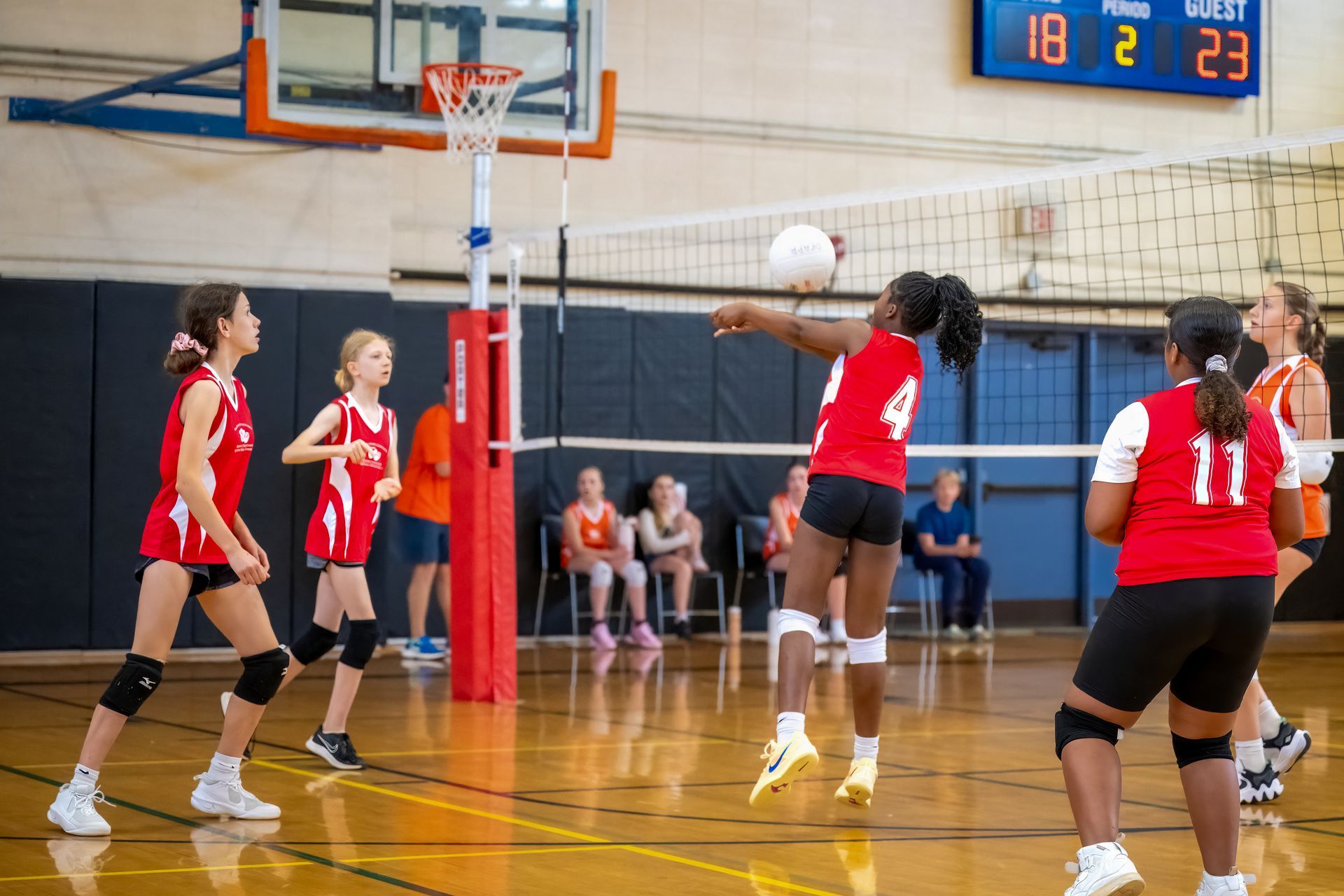 A volleyball player in a red jersey jumps to hit a ball over the net during a match in a gymnasium.