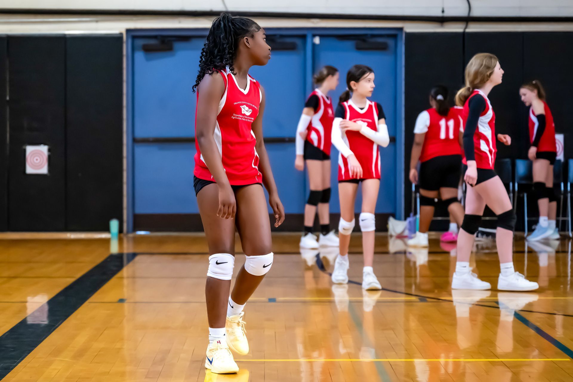 A group of players in red and white athletic uniforms standing on a hardwood basketball court in front of blue doors.