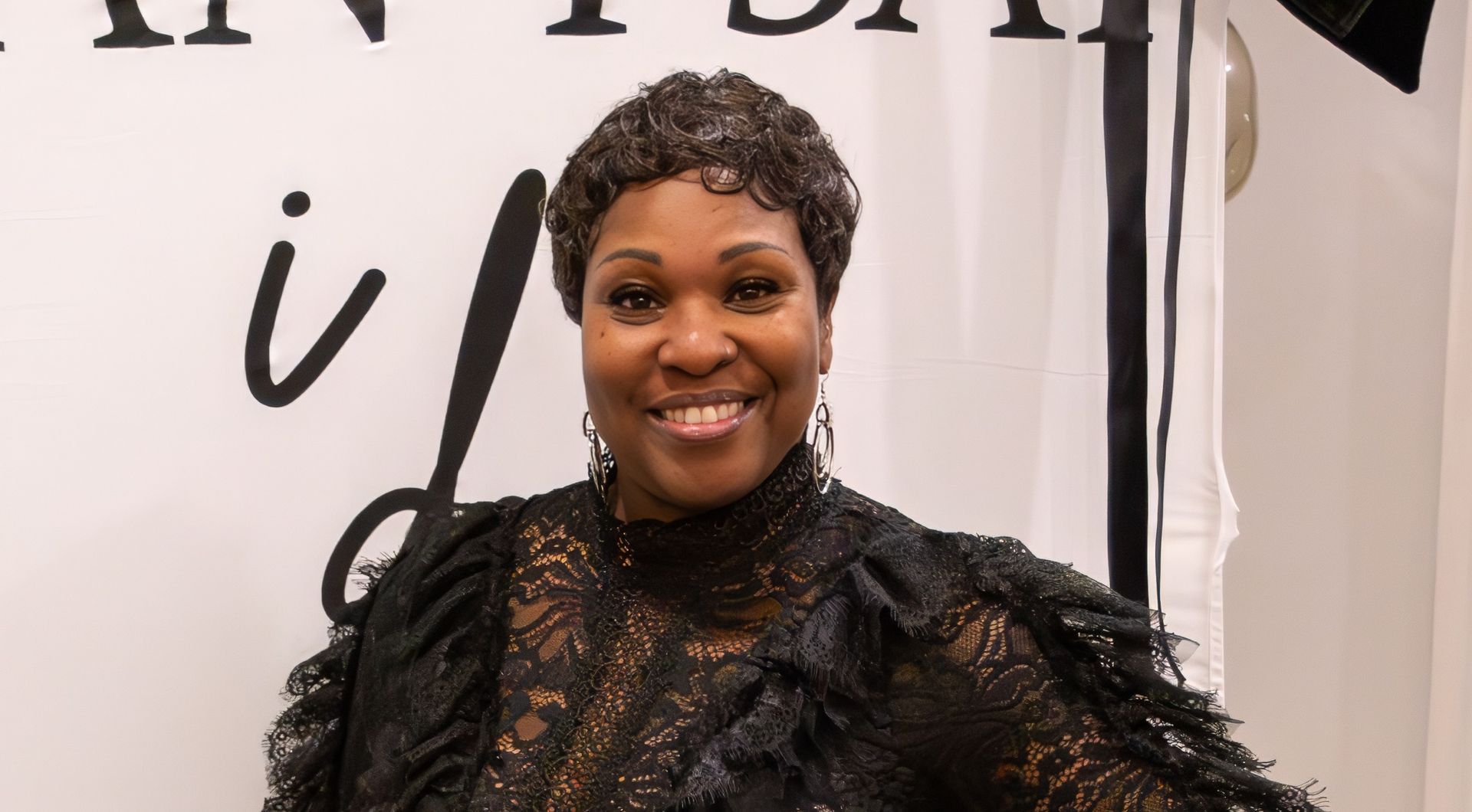 A smiling person with short, dark curly hair, wearing a black lace dress, stands in front of a white backdrop with text.