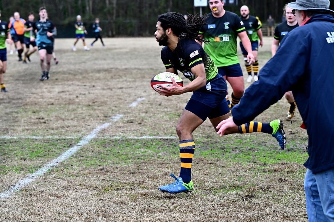 A rugby player in a dark jersey with yellow striped socks runs across a grass field holding a rugby ball.