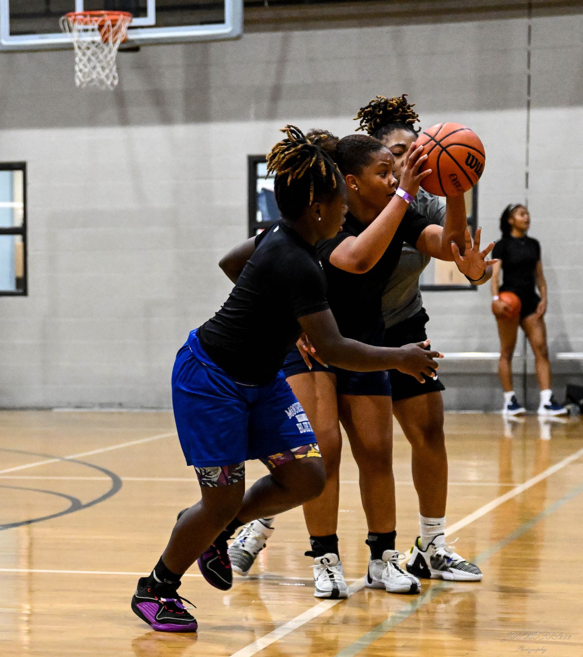 Three players in black shirts and athletic shorts practice basketball drills on an indoor court.