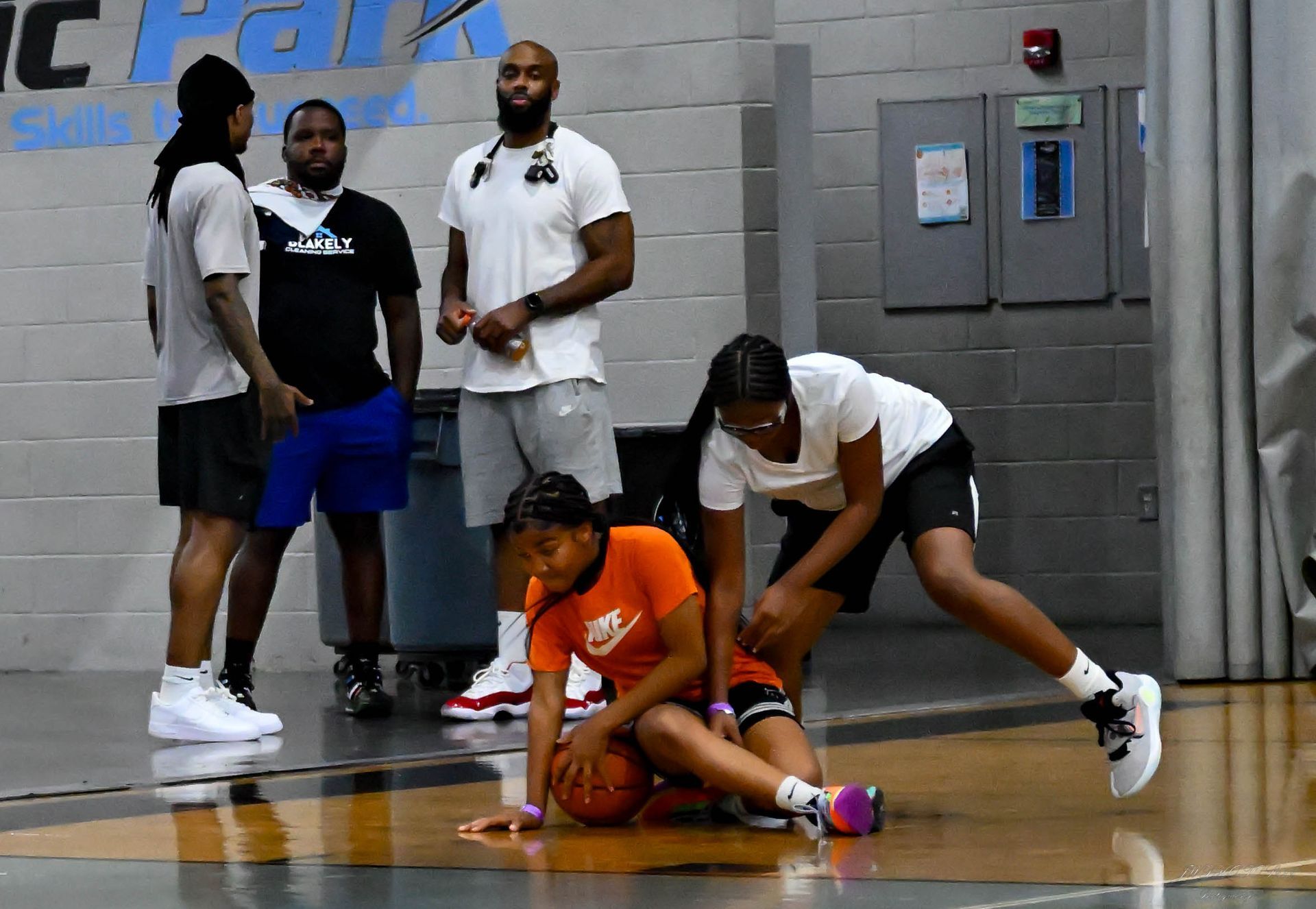 A basketball player in an orange shirt sits on the court with a basketball, while another player assists her.