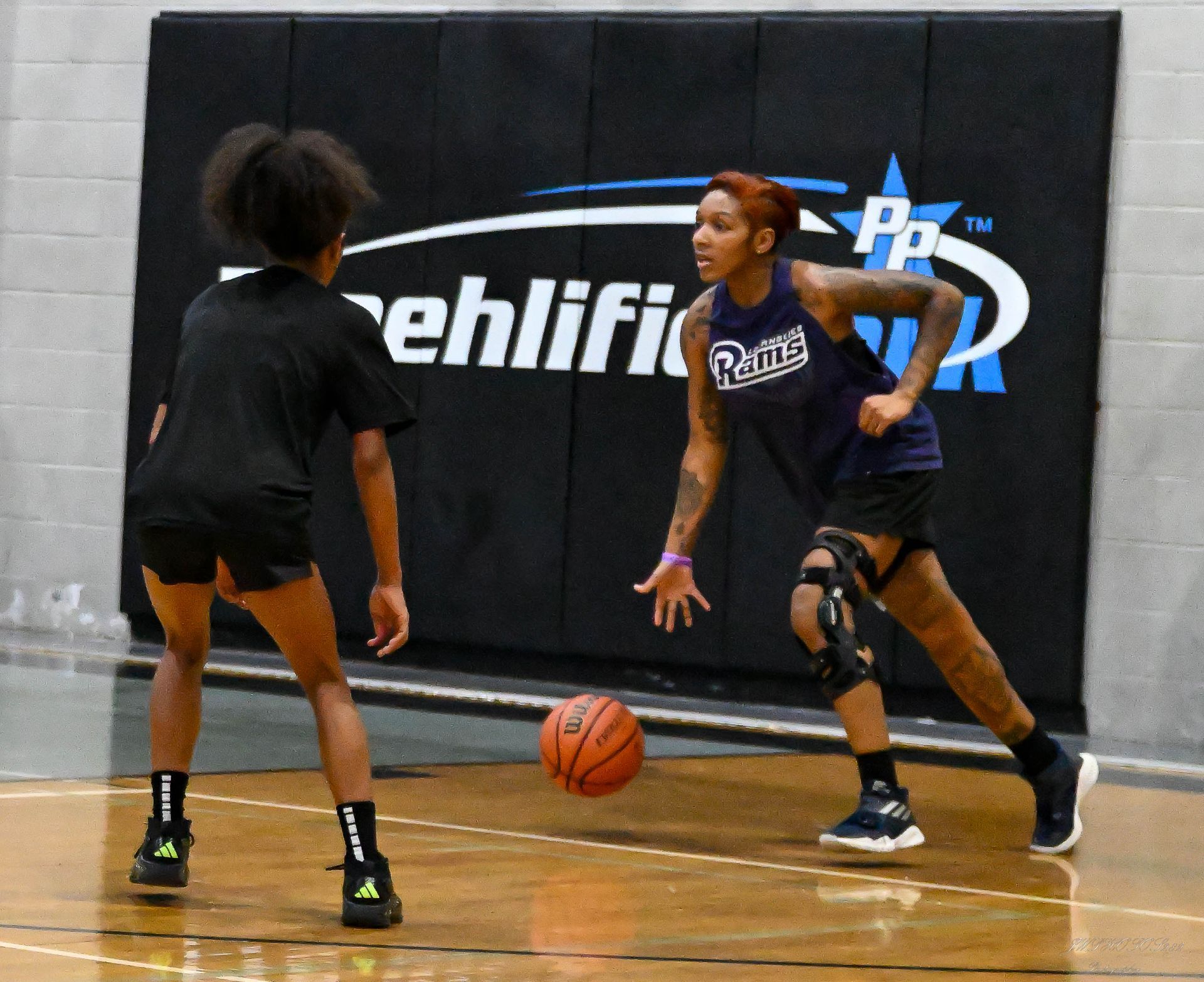 Two basketball players on an indoor court, one dribbling toward the other who is defending.