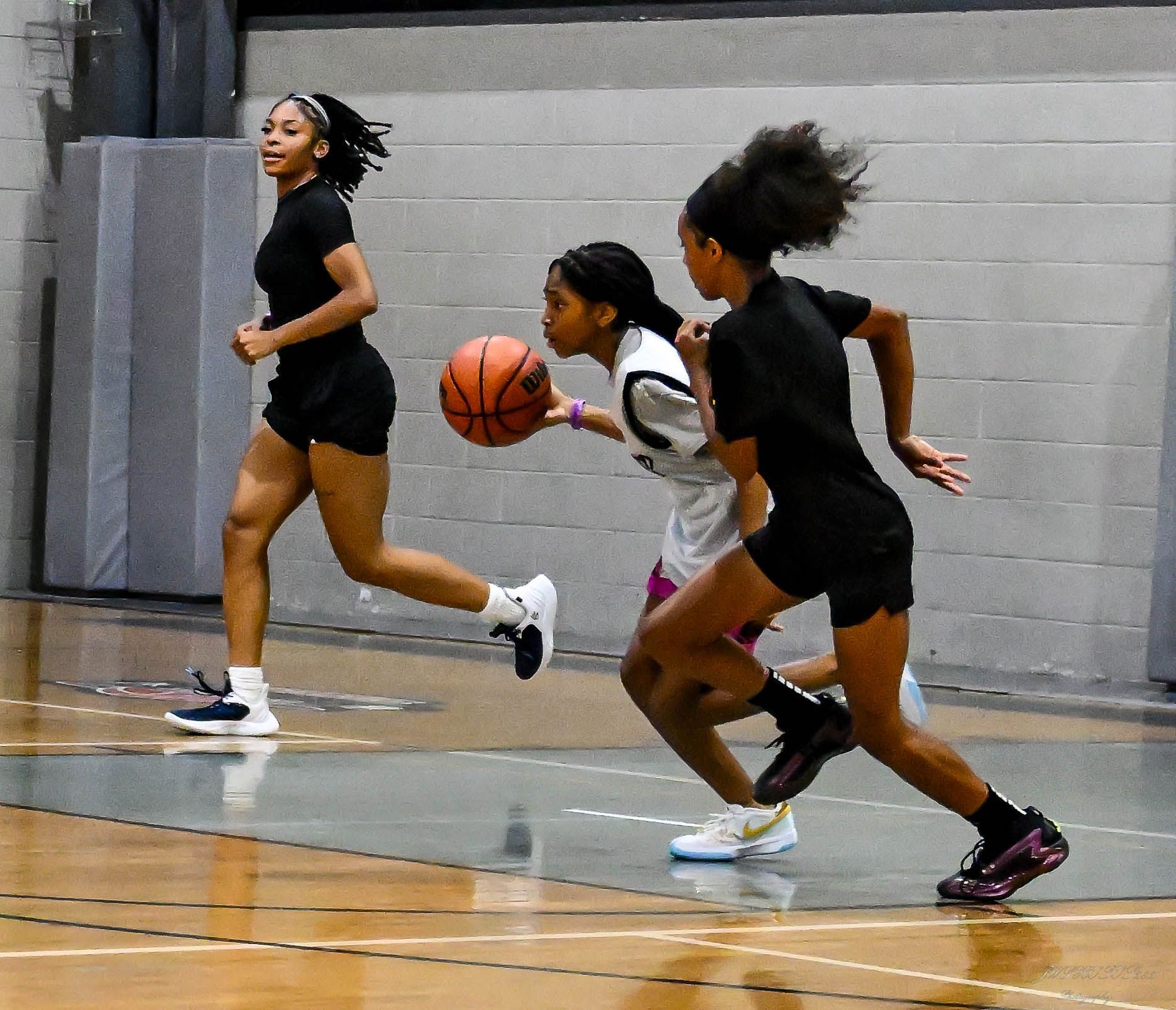 Three athletes run on an indoor basketball court, one dribbling a basketball while the others follow closely.