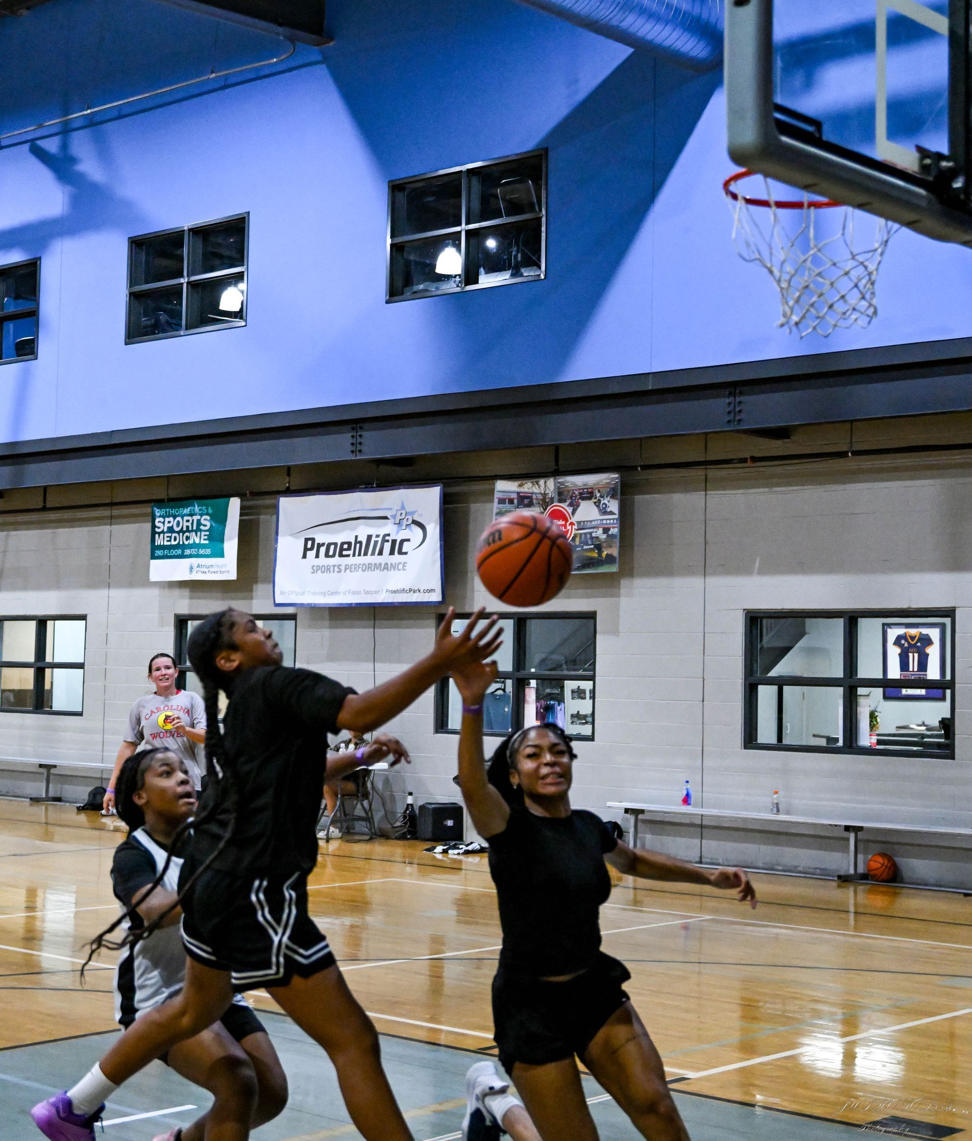 Three basketball players in black uniforms compete for a rebound under the hoop in an indoor court.