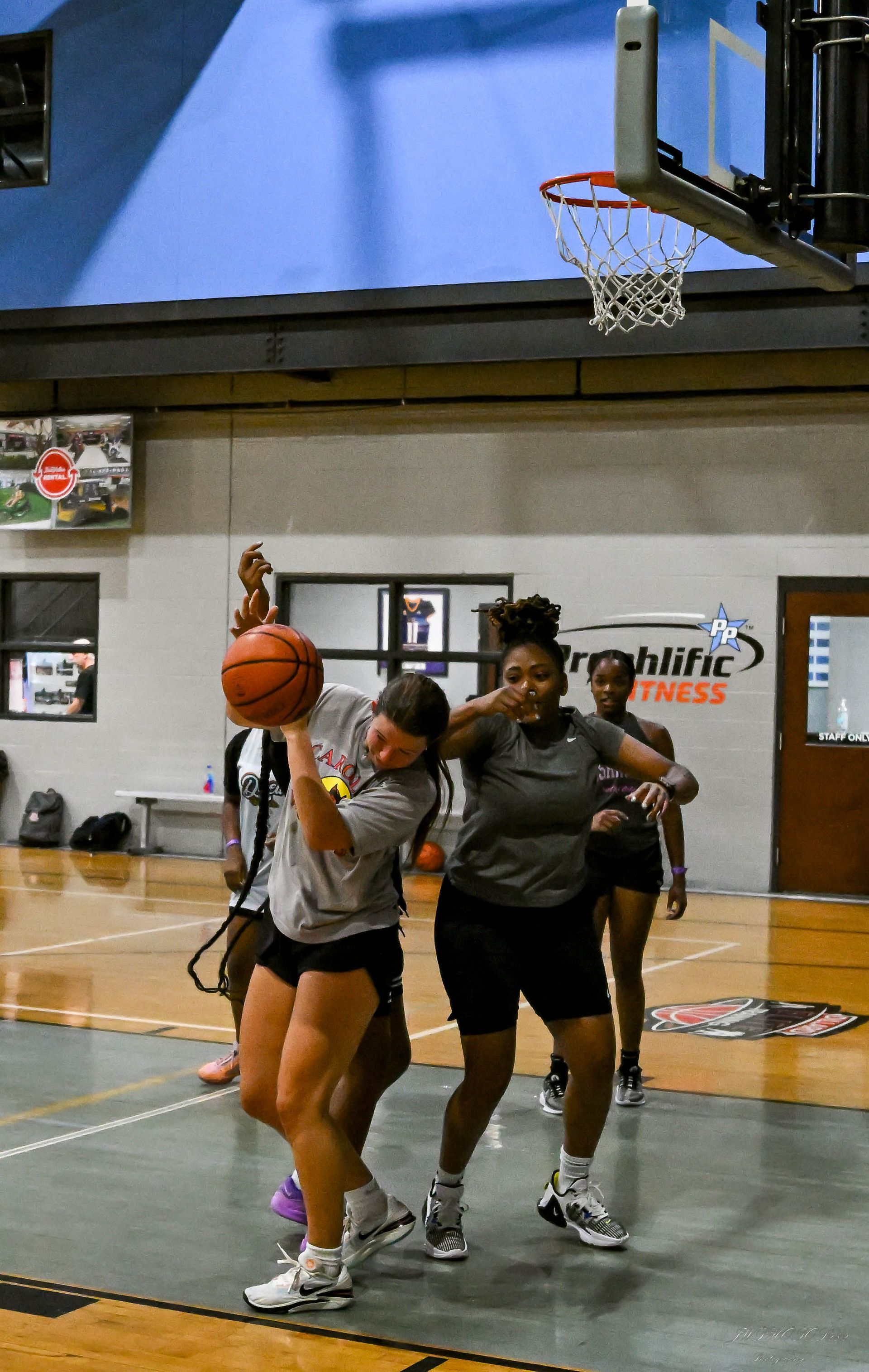 Three people practice basketball drills on an indoor court, with one athlete reaching up toward the hoop with the ball.