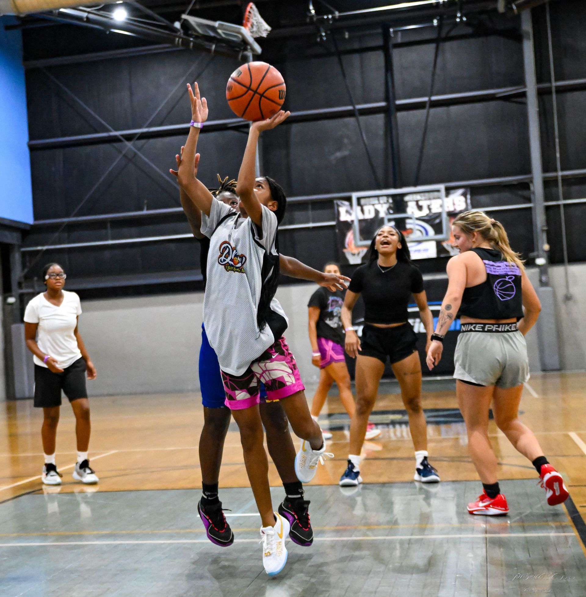 Basketball players in a gym, with one athlete jumping to shoot the ball while being defended.