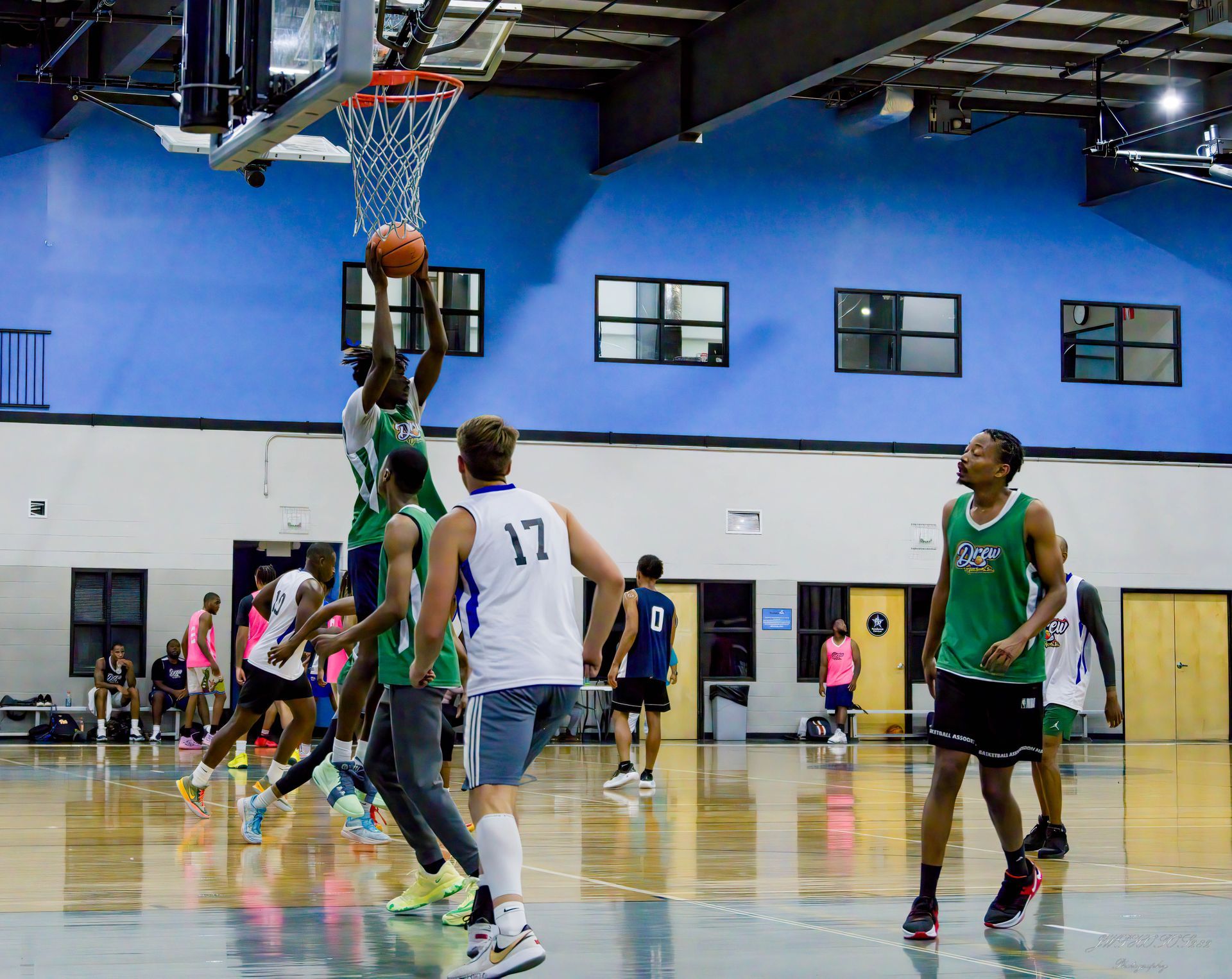 A basketball player in a green jersey jumps to score, with other players on an indoor court watching.
