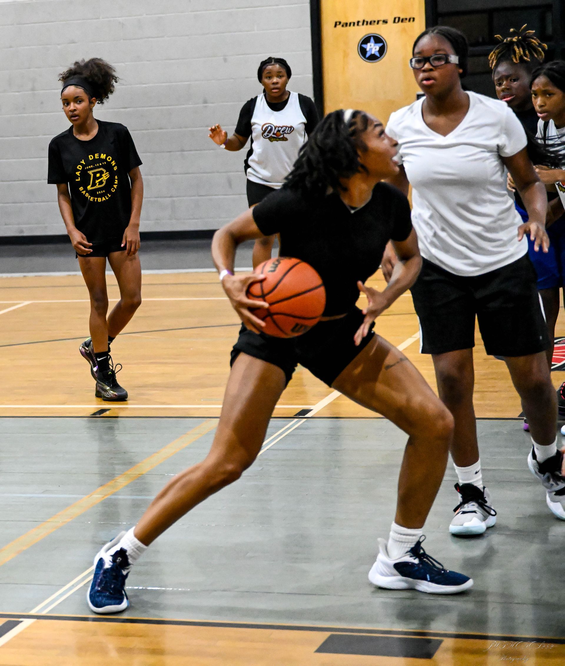 Players in athletic wear practice basketball drills on an indoor court, with one player dribbling as others watch.