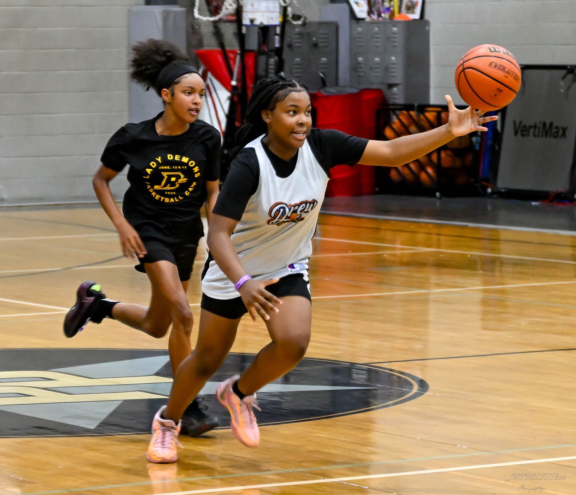 A player in a white jersey reaches for a basketball during a game while a teammate in a black shirt runs behind them.