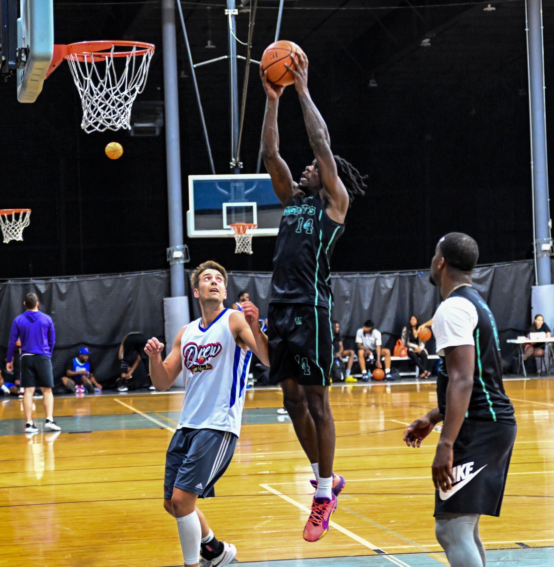 A basketball player in a black uniform jumps to dunk a ball, watched by two other players on an indoor court.
