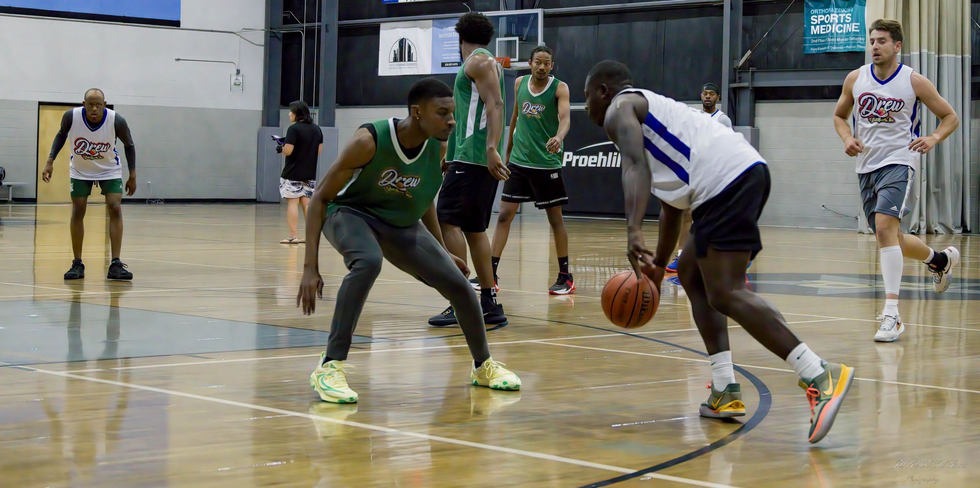 Basketball players in white and green jerseys compete on an indoor court, with one player dribbling the ball.
