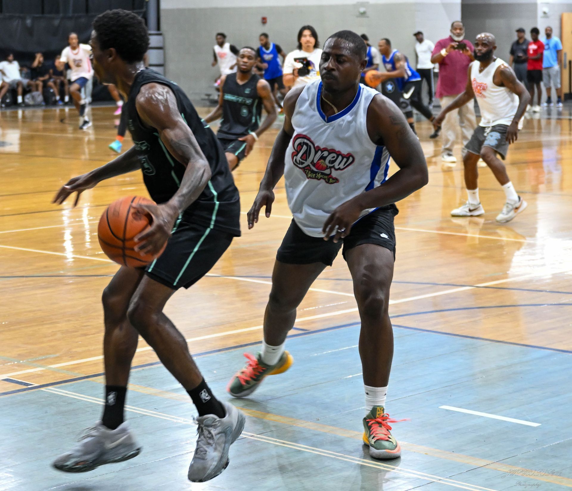 A basketball player in a black uniform dribbles against a defender in a white Drew League jersey on a hardwood court.