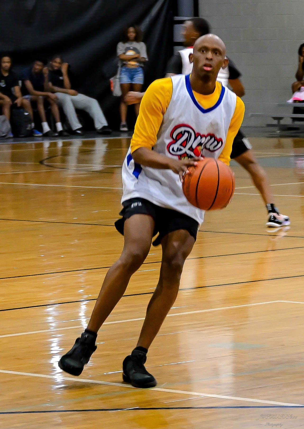 A person wearing a yellow and white jersey dribbles a basketball on an indoor wooden court.