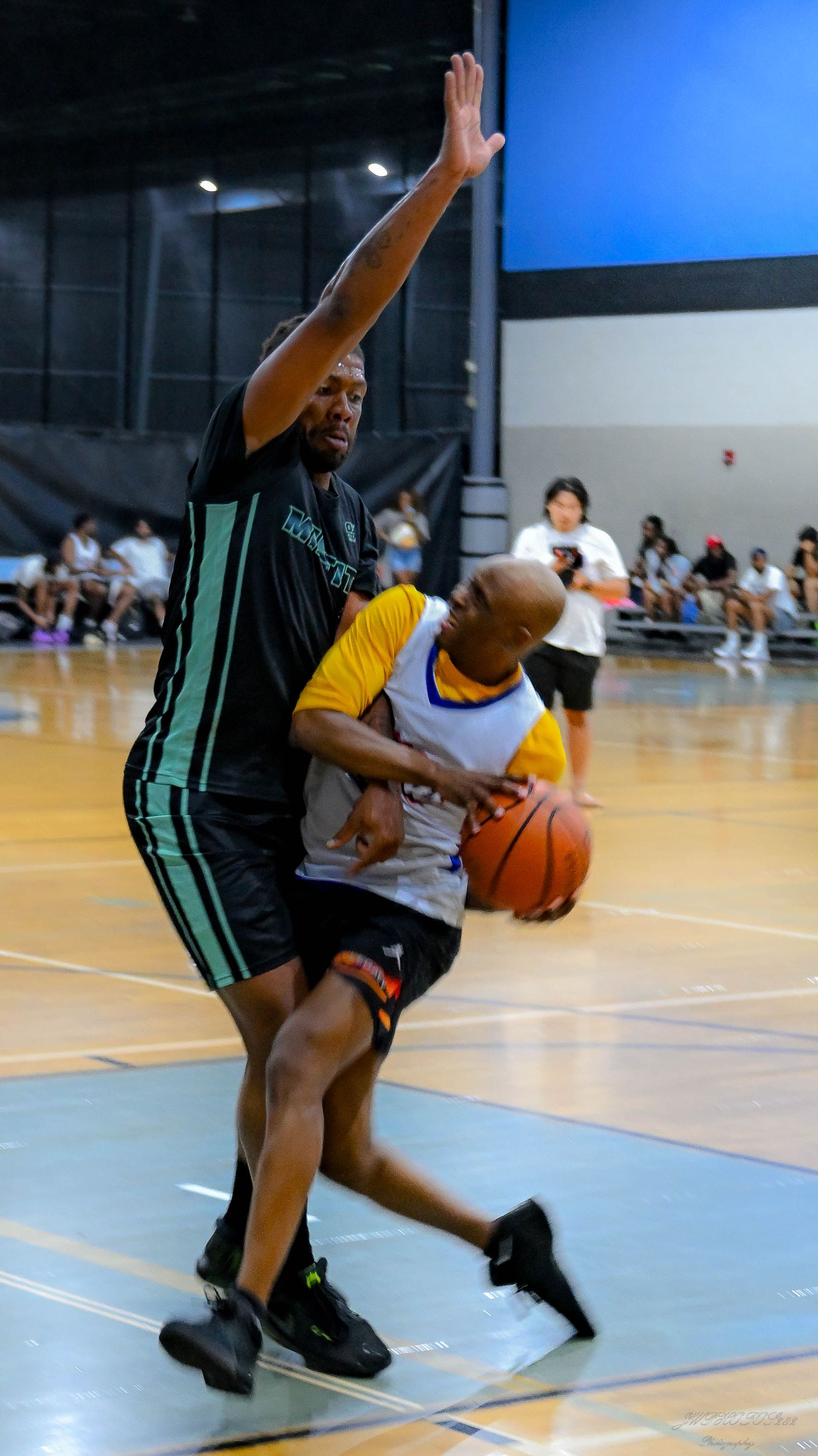 Two basketball players in a gym; one in a black uniform defends a player in a yellow and blue uniform holding the ball.