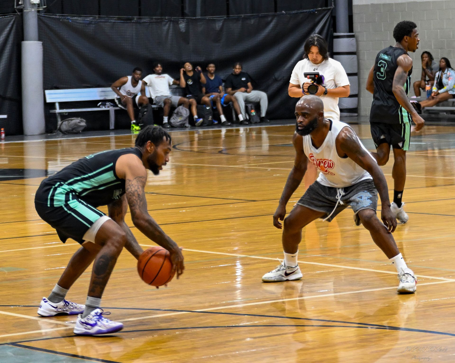 Two basketball players face each other on an indoor court, with one dribbling the ball while the other plays defense.