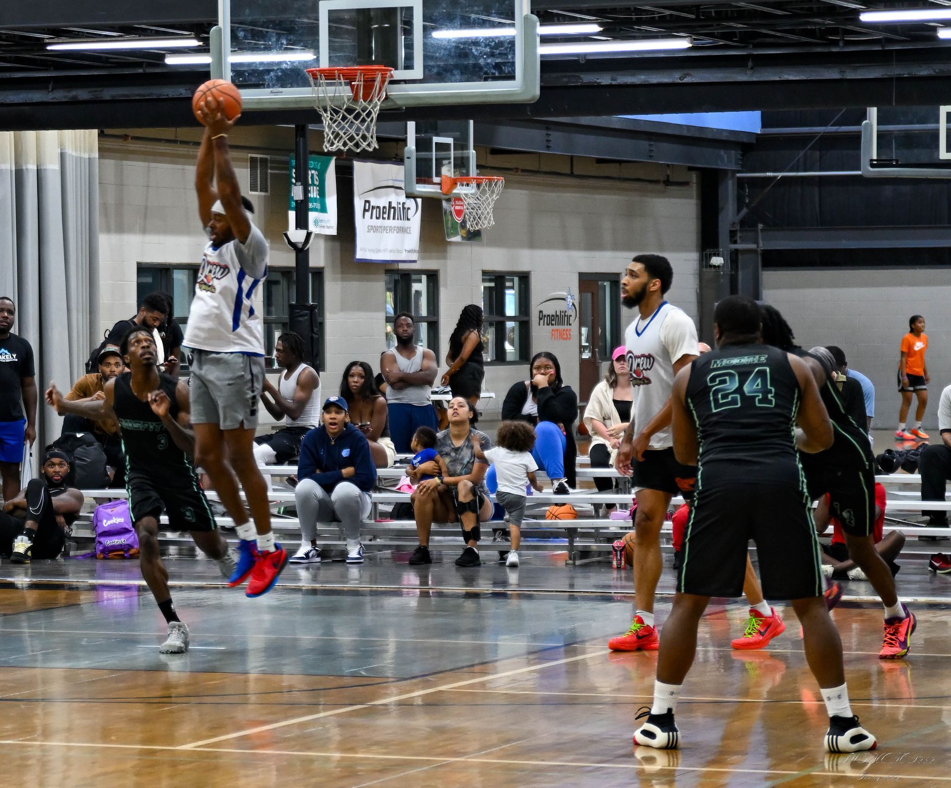 A basketball player jumps to shoot as others watch on an indoor court with spectators in the background.