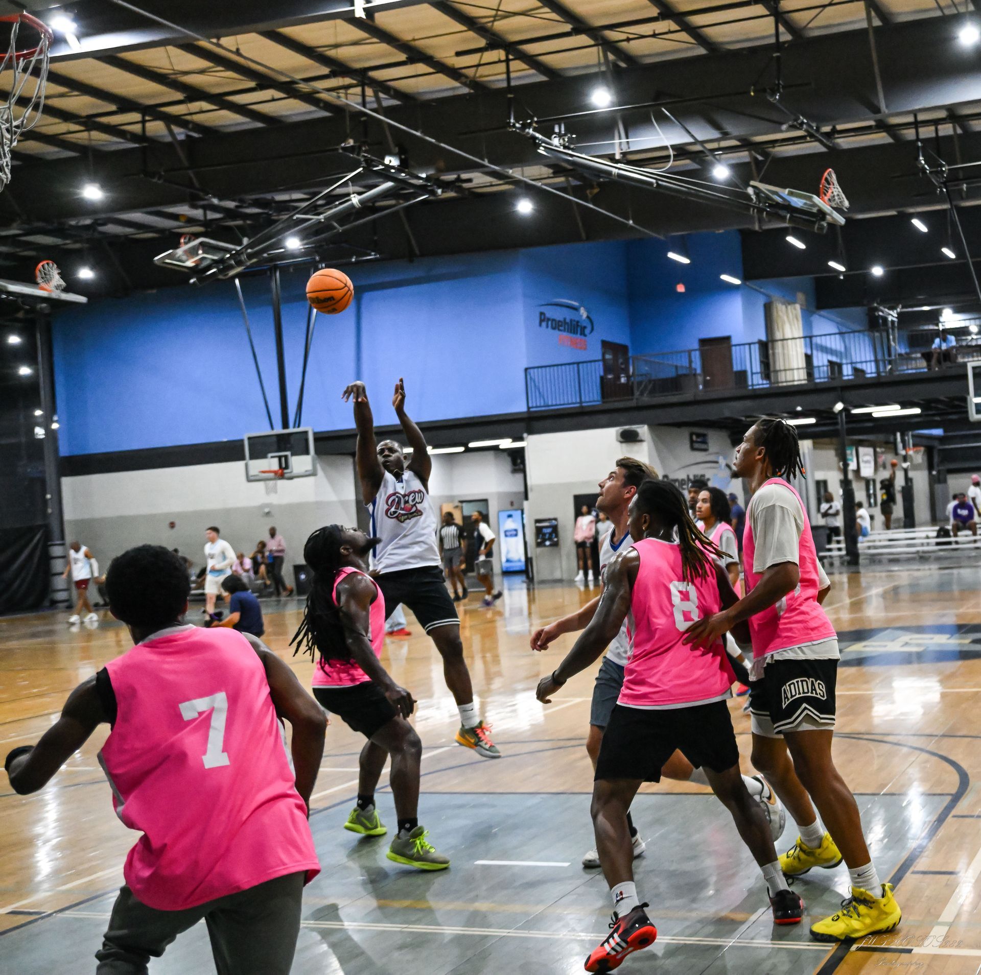 A basketball player leaps for a jump shot while teammates and opponents in pink jerseys watch on an indoor court.