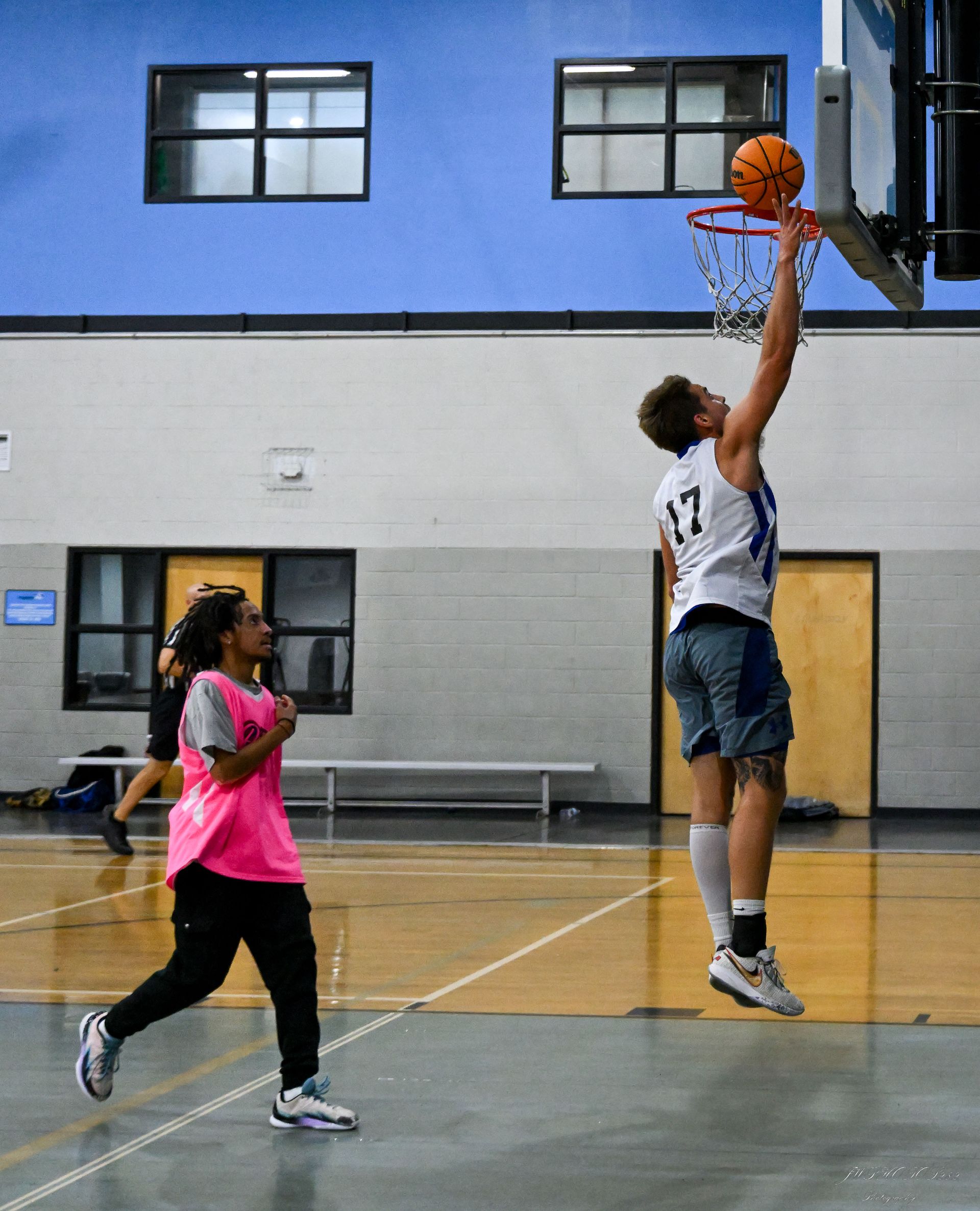 A player in a white jersey shoots a basketball into the hoop while a player in a pink jersey watches on a gym court.
