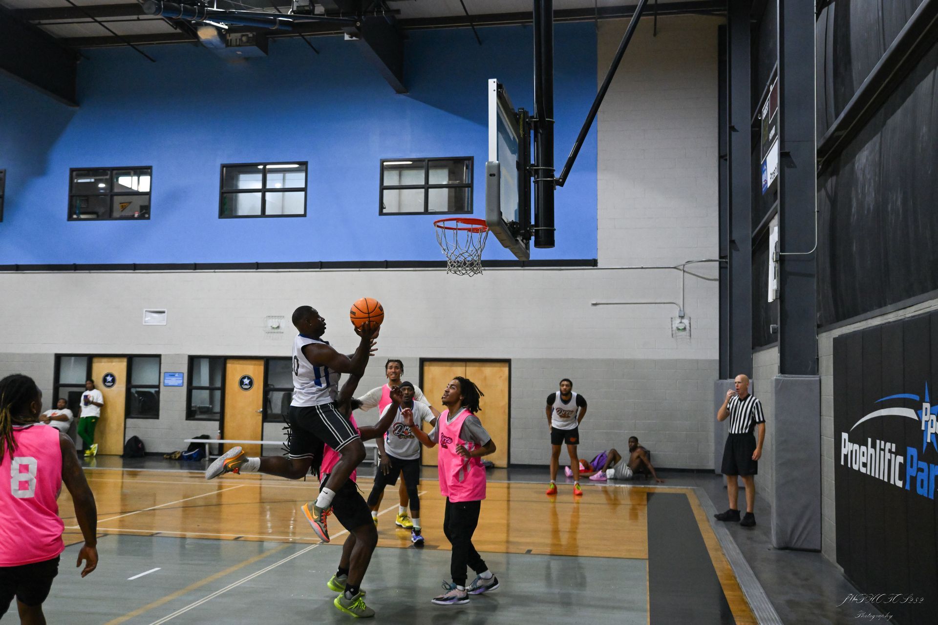 A basketball player in a white jersey leaps for a shot, surrounded by players in pink and white jerseys on an indoor court.