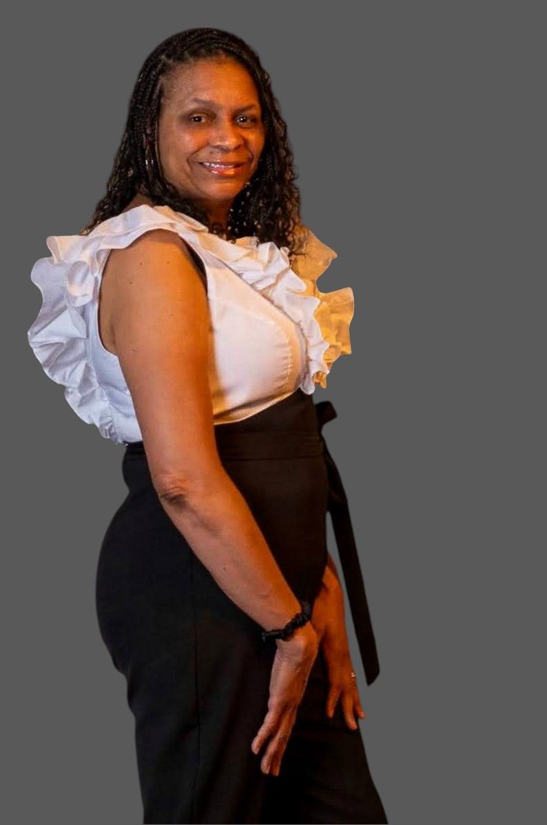A person with shoulder-length curly dark hair smiles, wearing a white ruffle-sleeved top and black high-waisted trousers.