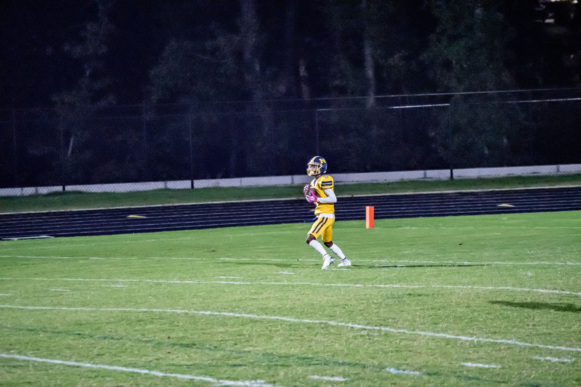 A football player in a yellow and black uniform runs across a green field at night, holding a football.
