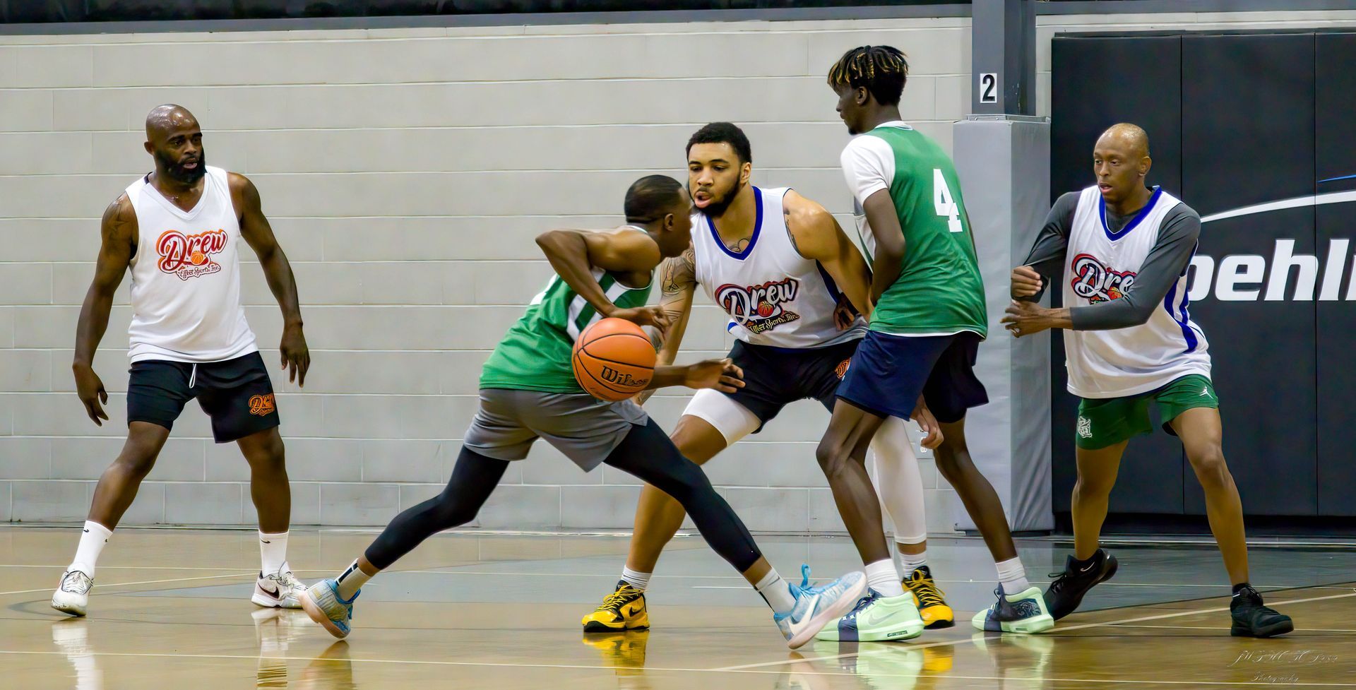 Basketball players in white and green jerseys practice on an indoor court, focusing on ball handling during a drill.