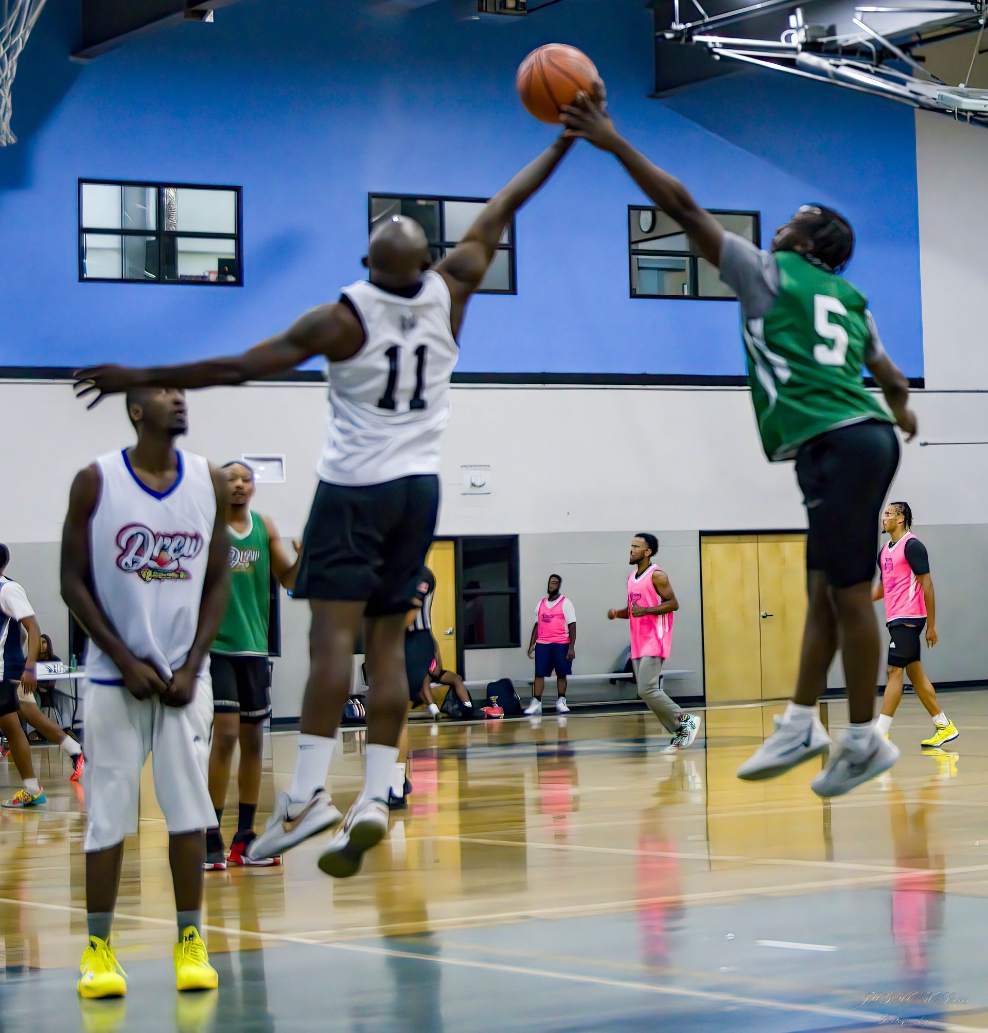 Two basketball players jump to contest a ball in an indoor gym, while other players stand on the court nearby.