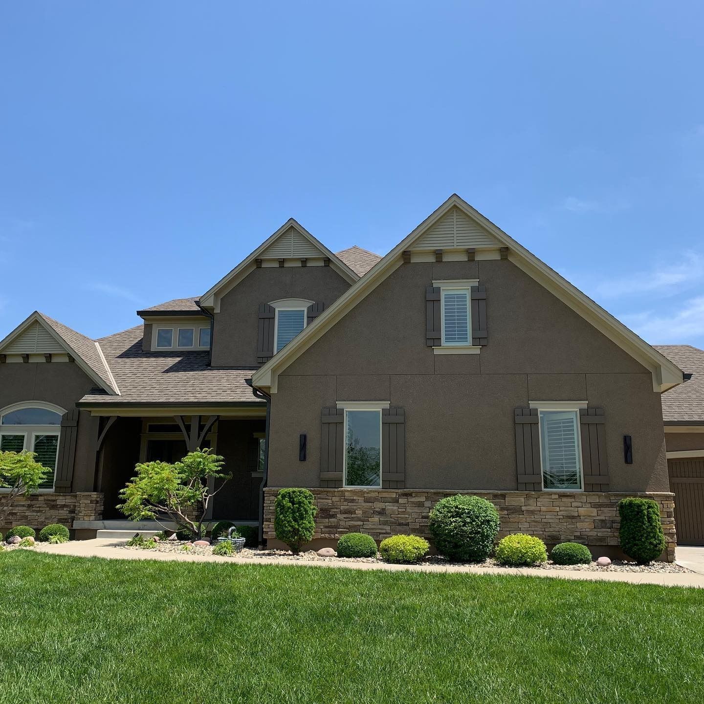 A tan, multi-gabled house with stone skirting, dark shutters, and a manicured lawn under a clear blue sky.