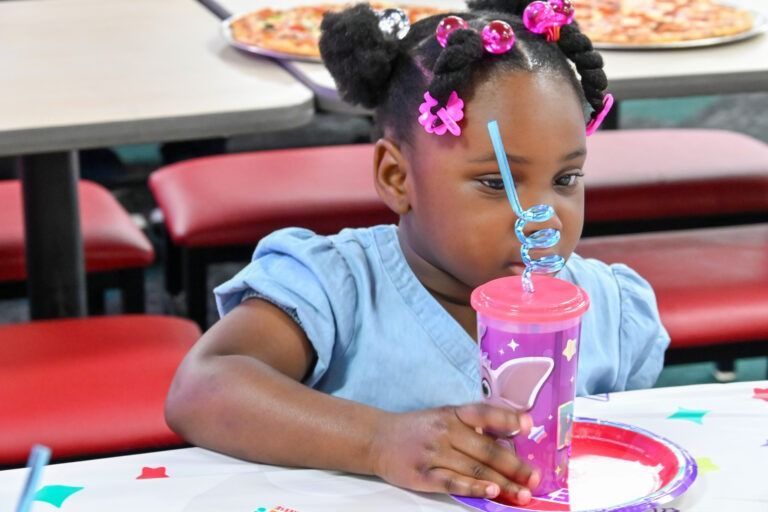 A child with hair in buns sits at a table, holding a purple cup with a curly blue straw, with pizza visible in the back.