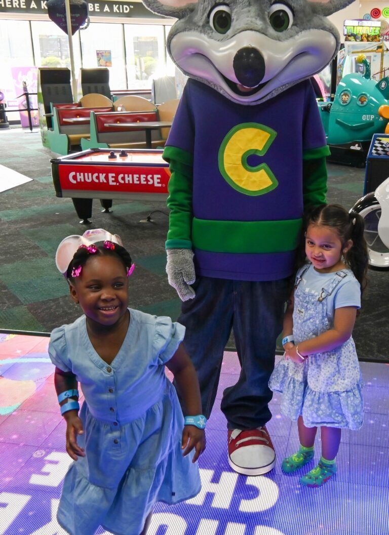 Two children standing on a colorful floor at a Chuck E. Cheese play center, posed with the mascot character.