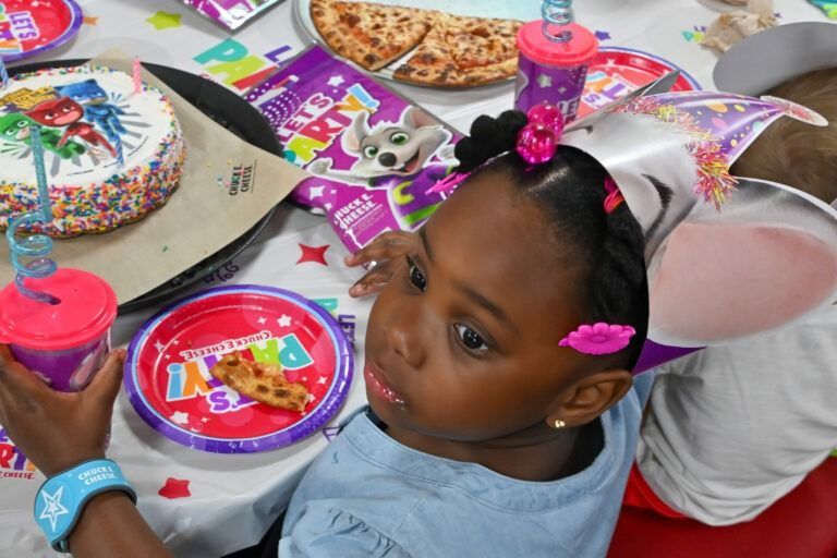 A child wears a party hat at a table with cake, pizza, and party-themed plates.