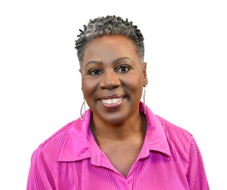 Smiling person with short, dark, textured hair wearing a vibrant pink collared shirt and long silver earrings.
