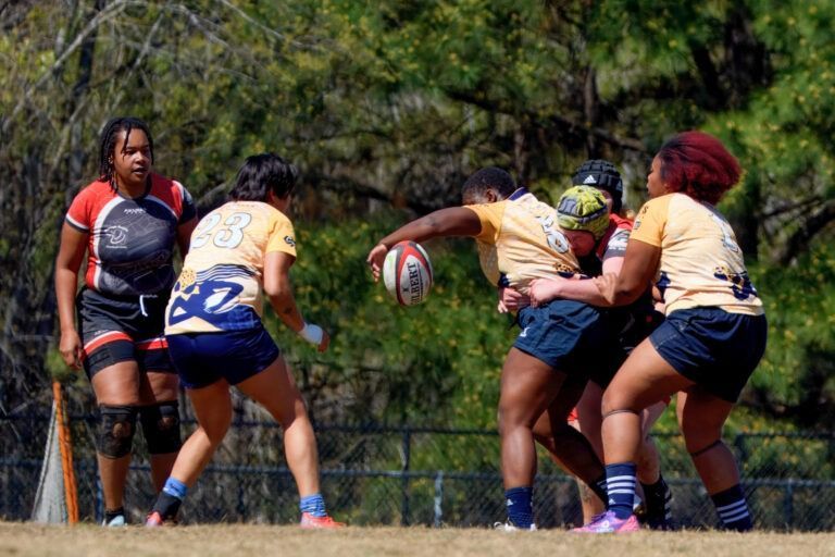 Rugby players in yellow jerseys pass a ball during a game on a sunny field with trees in the background.