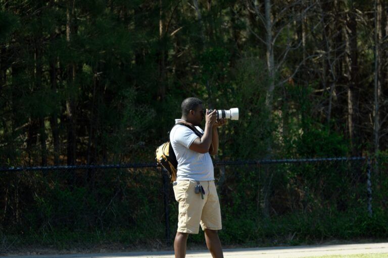 A person wearing a light-colored shirt and khaki shorts stands outdoors, using a camera with a long white lens.