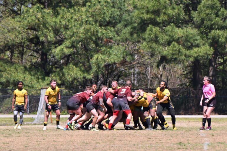 Players in gold and maroon jerseys contest a rugby scrum on a grass field, while an official in pink watches nearby.