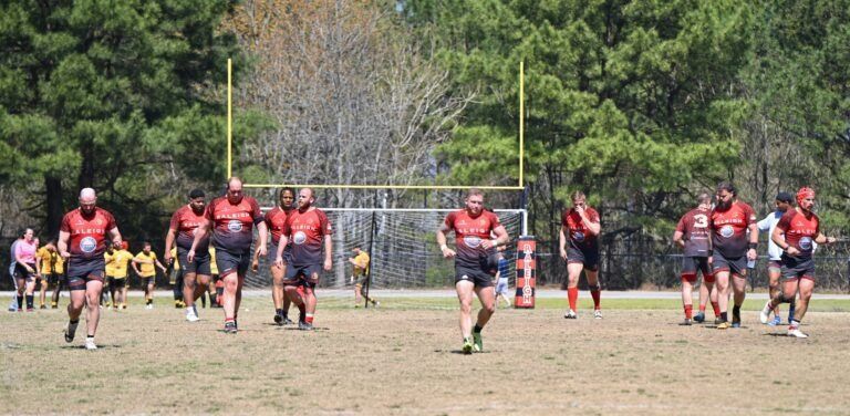A team of rugby players in matching red and black uniforms walk across a grass field near goalposts.