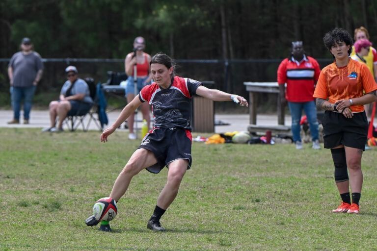 A player kicks a rugby ball on a grassy field, observed by a referee and spectators in the background.