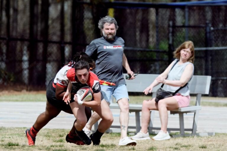 A rugby player in a red and black uniform runs with the ball while being tackled, near a bystander and a seated spectator.