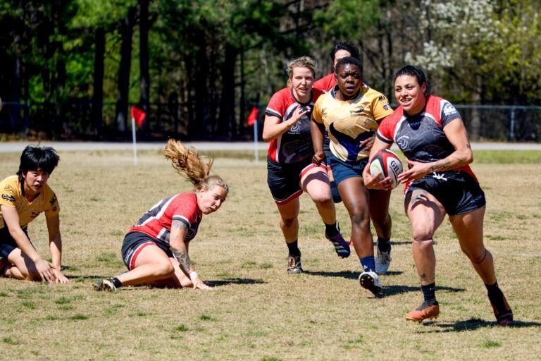 A group of rugby players in athletic uniforms on a grassy field; one player runs forward while others pursue or fall.