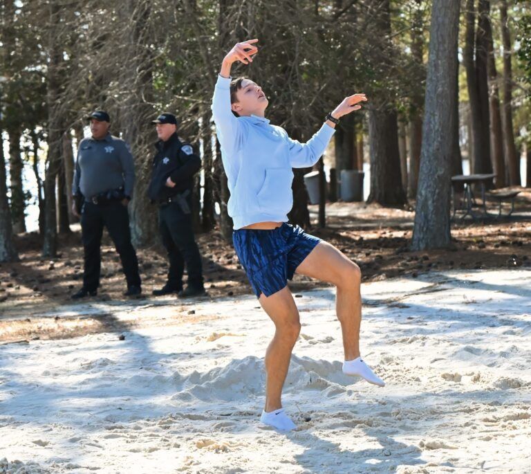 A person in a light blue hoodie and dark shorts balances on one foot on a sandy surface, with two figures in uniform behind.