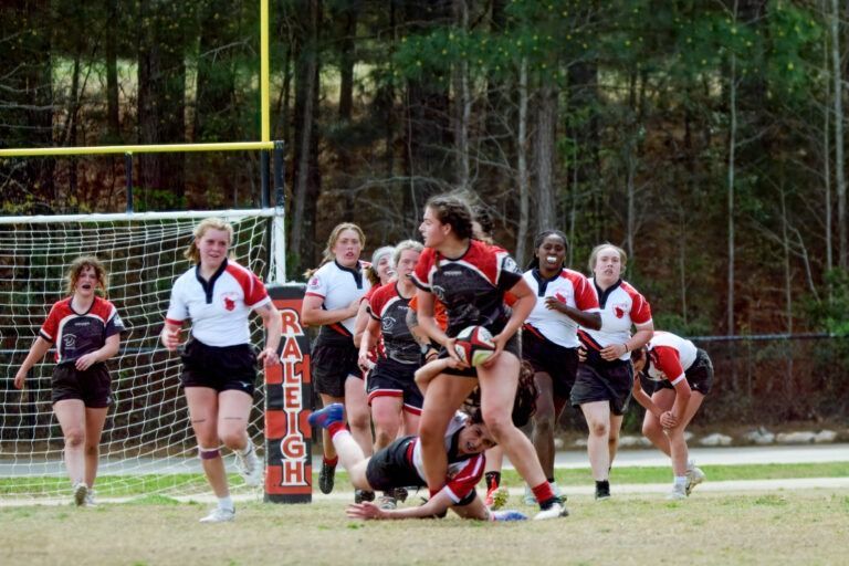 Players in red and white rugby uniforms compete for the ball on a grassy field in front of a goalpost.