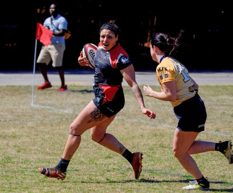 A player in a black and red uniform runs with a rugby ball while pursued by an opponent in a yellow and black jersey.