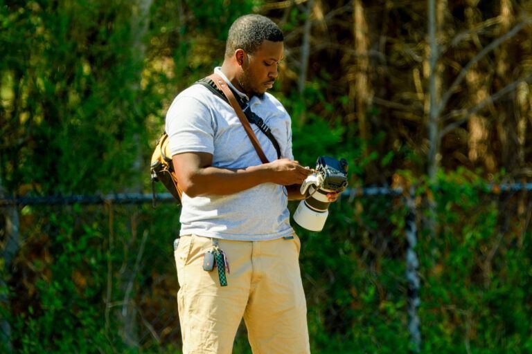 A person wearing a light polo and tan pants adjusts camera equipment while standing in front of a chain-link fence.