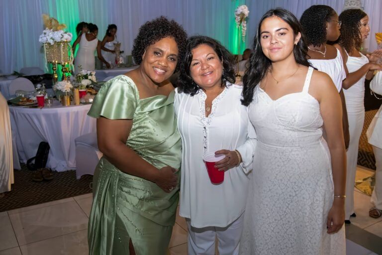 Three people smile together at a formal event, with the person on the left in a green dress and two in white outfits.
