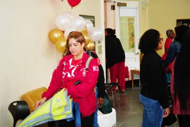 A group of people gathers in a room with gold and white balloons, one person holding a folded yellow umbrella.