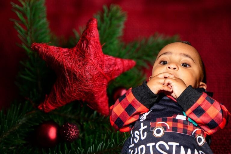 A baby in plaid pajamas looks upward next to a large red star and evergreen branches with holiday ornaments.