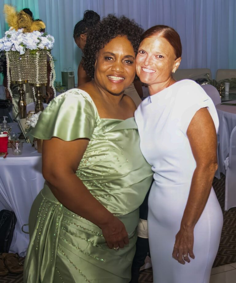 Two people smiling, standing side-by-side in a room with table settings, one in a pale green dress and one in white.