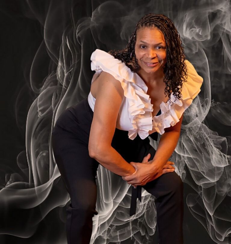 A person wearing a white ruffled top and black pants leans forward against a dark background with swirling white smoke.