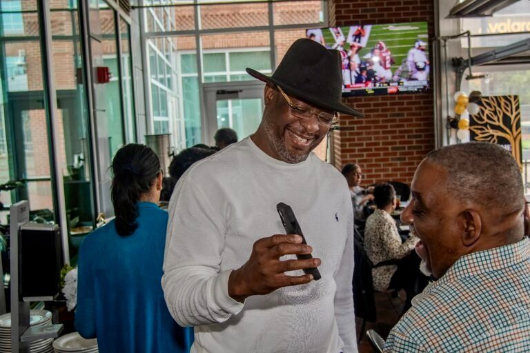 A person in a black fedora and white sweater shows a smartphone to another person at an indoor gathering.